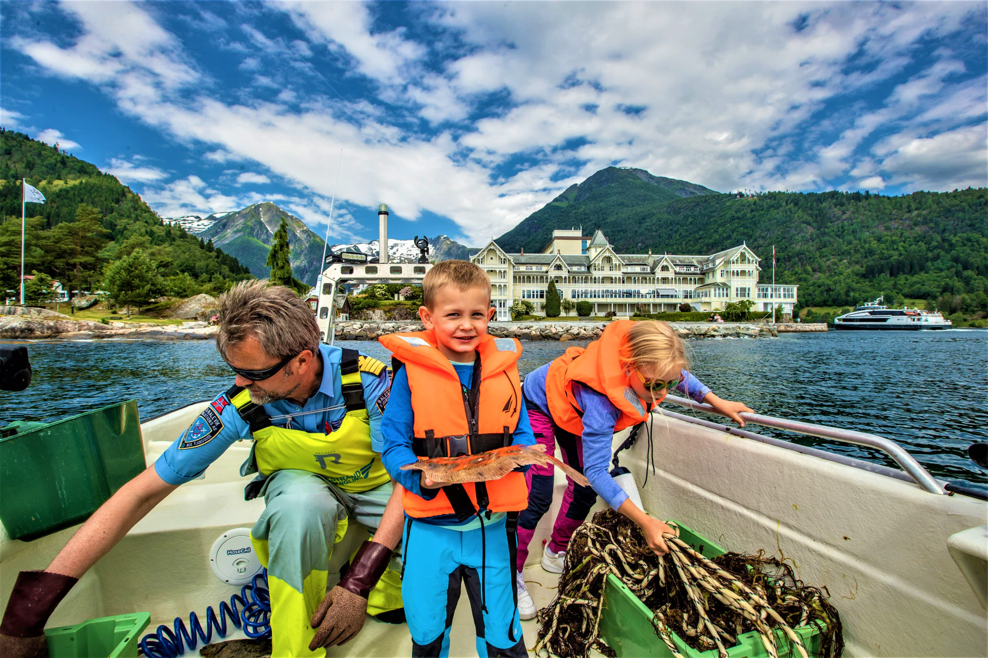 "Captain Tor of Balestrand Fjord Angling teaching children traditional gillnet fishing on the Sognefjord. A young boy proudly holds a fresh flatfish (plaice) while a girl explores the fishing net. The historic Kviknes Hotel and majestic mountains of Balestrand, Norway, provide a stunning backdrop. Highlighting the 'Fiskeskole' educational experience and authentic family fjord adventures."