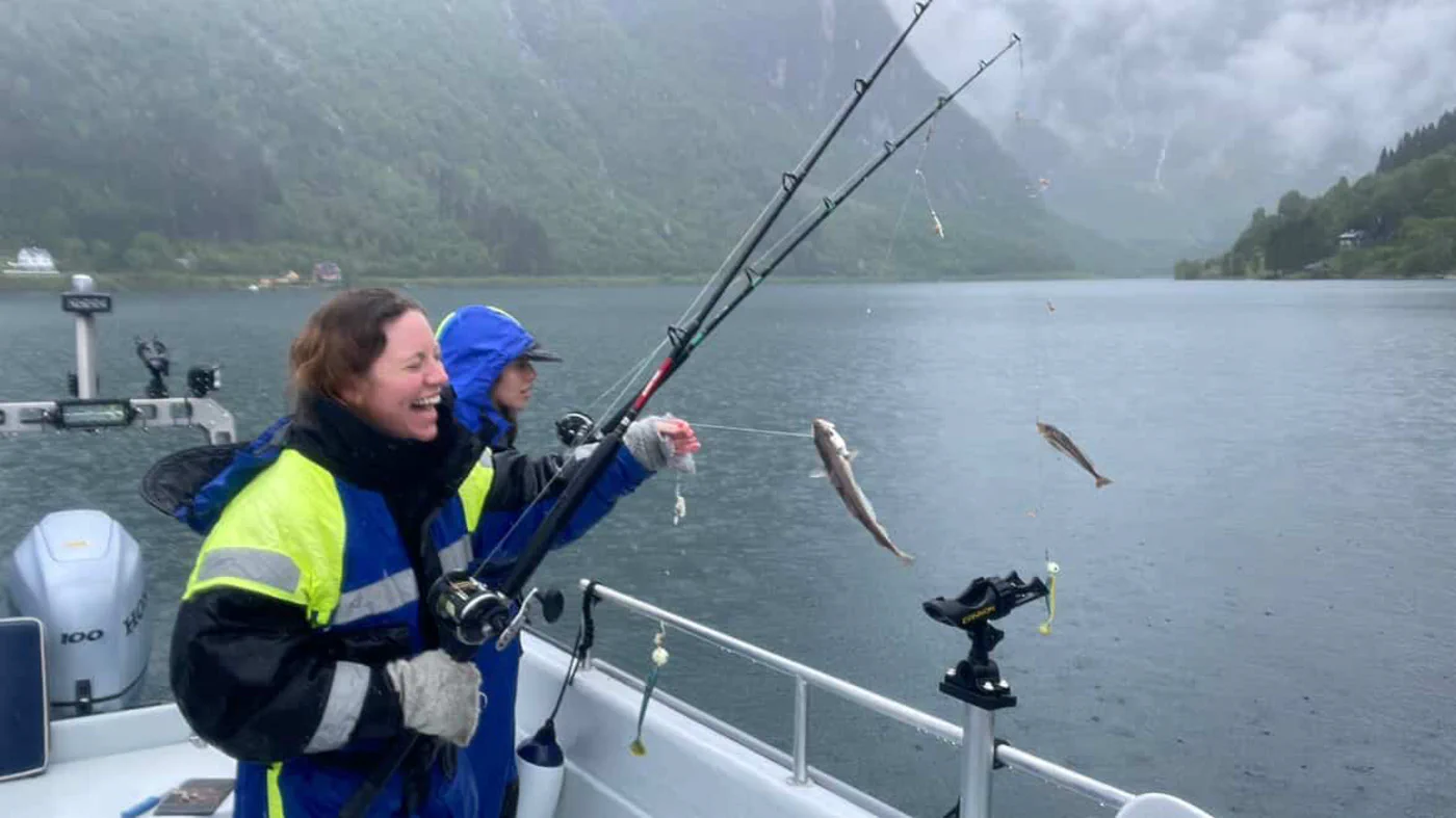 "Two Italian women from New York City experiencing the raw beauty of a rainy day on the Sognefjord with Balestrand Fjord Angling. One woman is laughing with pure joy while reeling in a fresh catch (whiting) on a fishing rod. Authentic Norwegian nature experience with misty mountains and rain, showcasing that 'Simplicity Creates Emotions' regardless of the weather. Highlighting adventure travel, female empowerment in the outdoors, and the 'Human Edge' in Balestrand, Norway."