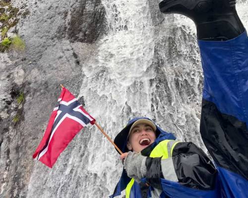 "A happy female guest smiling on a guided boat trip with Balestrand Fjord Angling in the Sognefjord, Norway. She is holding a Norwegian flag, wearing professional flotation gear and boots, with a powerful waterfall in the background. The scene showcases the pure joy of 'Simplicity Creates Emotions' and authentic 'Human Edge' experiences in Balestrand, Norway."