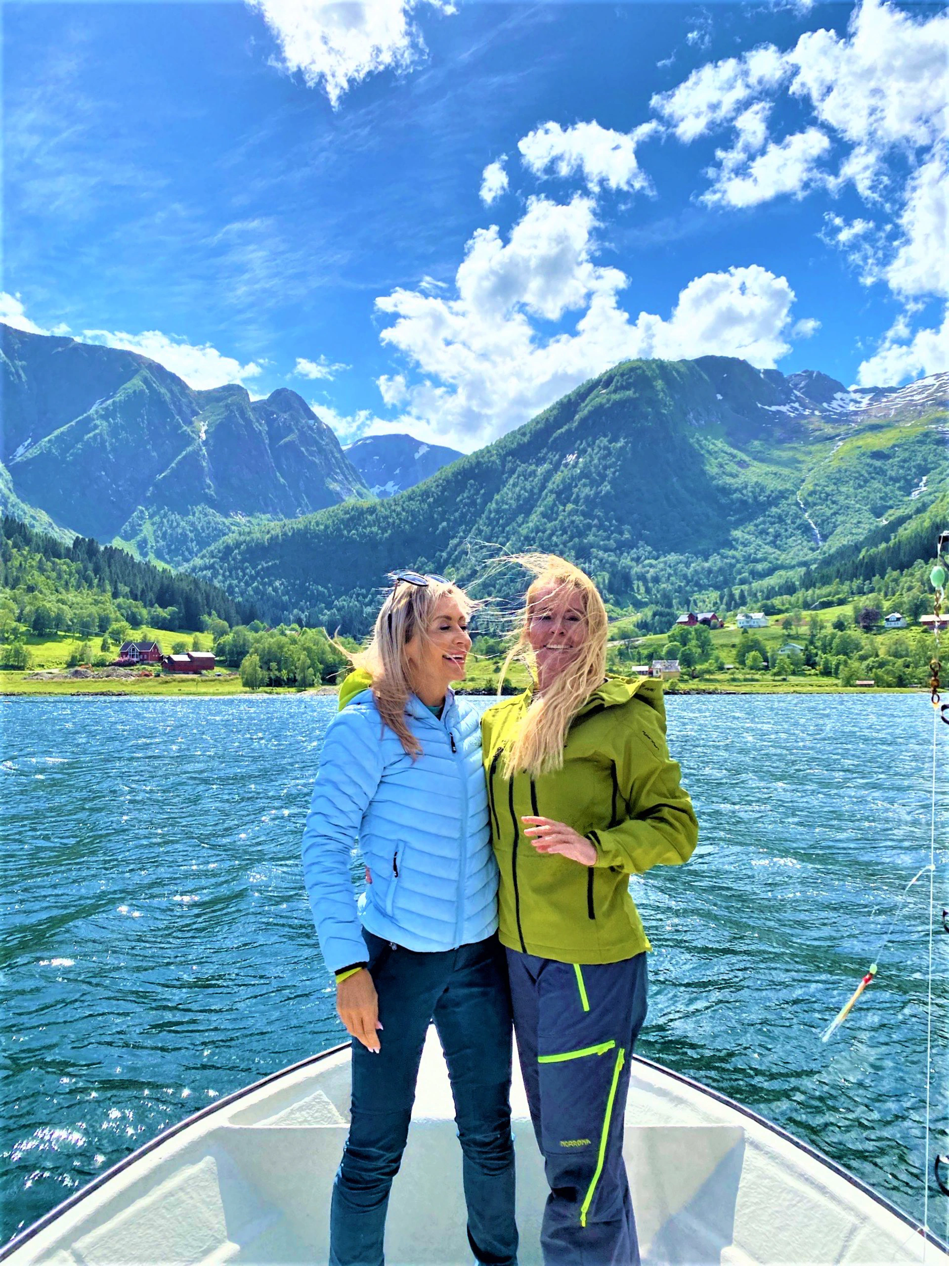 "Two smiling and energetic women in hiking clothes stand close together and laugh on board a boat in the Sognefjord. Behind them rise green mountain slopes with snow-capped peaks and waterfalls under a blue sky with white clouds."