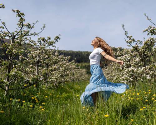 Woman dancing joyfully in a blossoming apple orchard in Balestrand, Sognefjord. The image captures the Norwegian 'springfulness' feeling, mental wellness, and authentic nature experience.