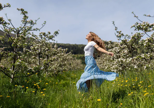 Woman dancing joyfully in a blossoming apple orchard in Balestrand, Sognefjord. The image captures the Norwegian 'springfulness' feeling, mental wellness, and authentic nature experience.