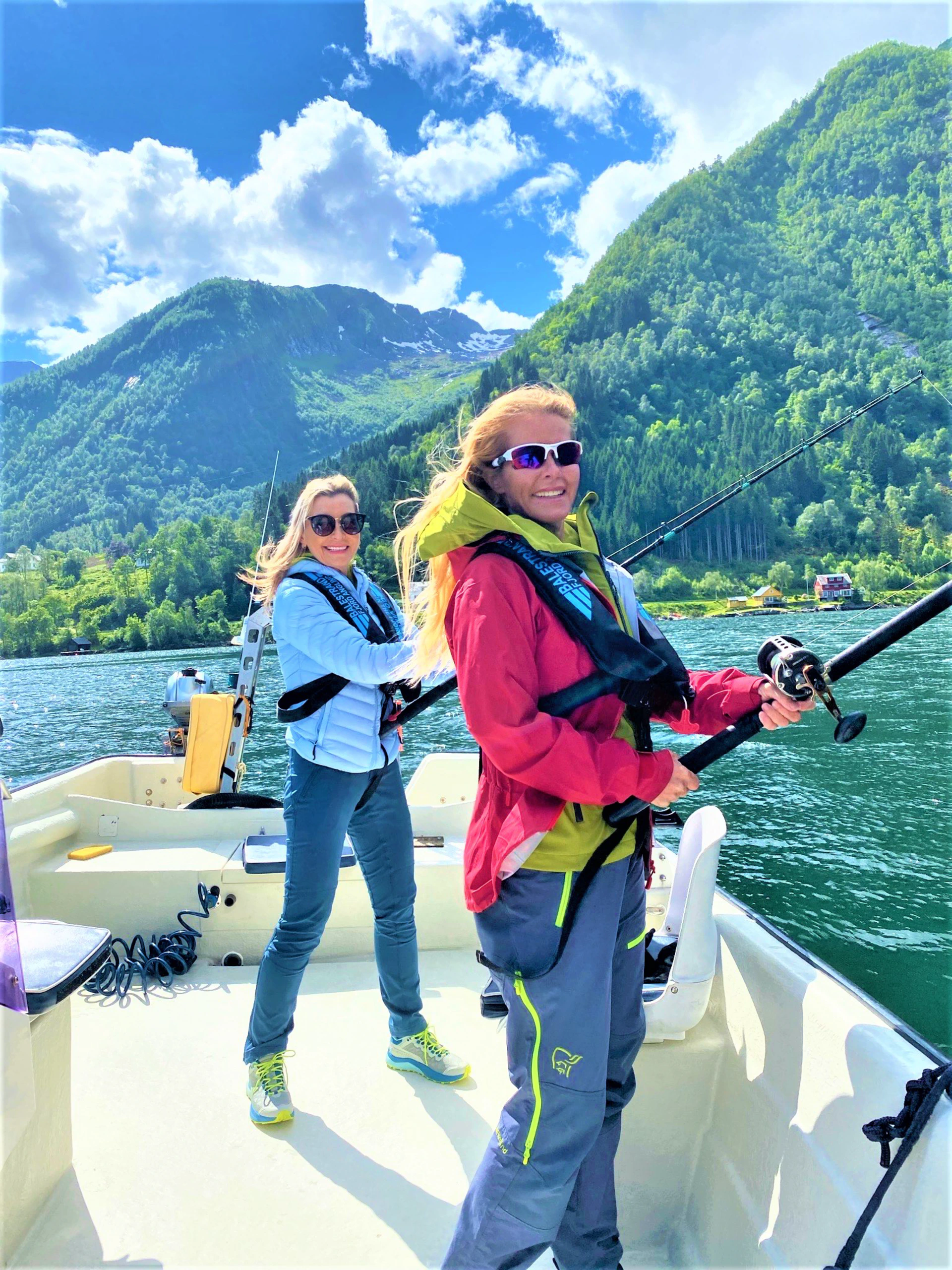 Two smiling women in life vests are fishing from a boat on a sunny day in a Norwegian fjord with steep green mountains in the background.